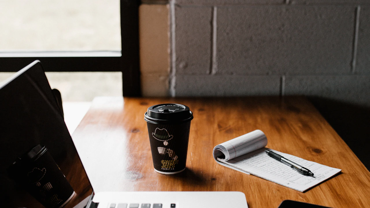 A laptop, notebook and coffee on a wooden table by a window.