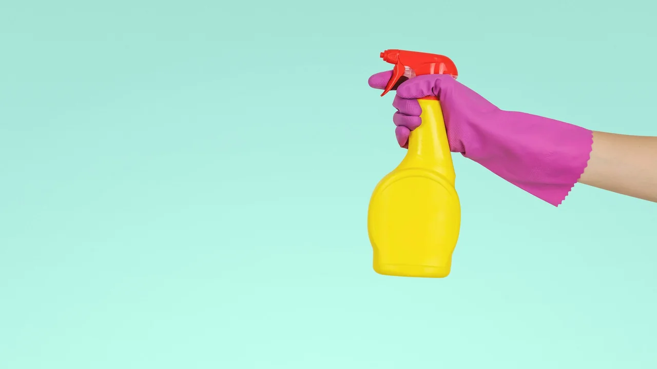 Gloved hand holding a yellow cleaning spray bottle against a mint background